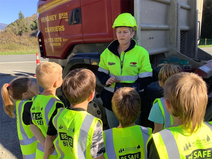 6 school children listen to and adult who is talking to them about forestry. They are all wearing hi-visibility vests and stand in front of a forestry truck